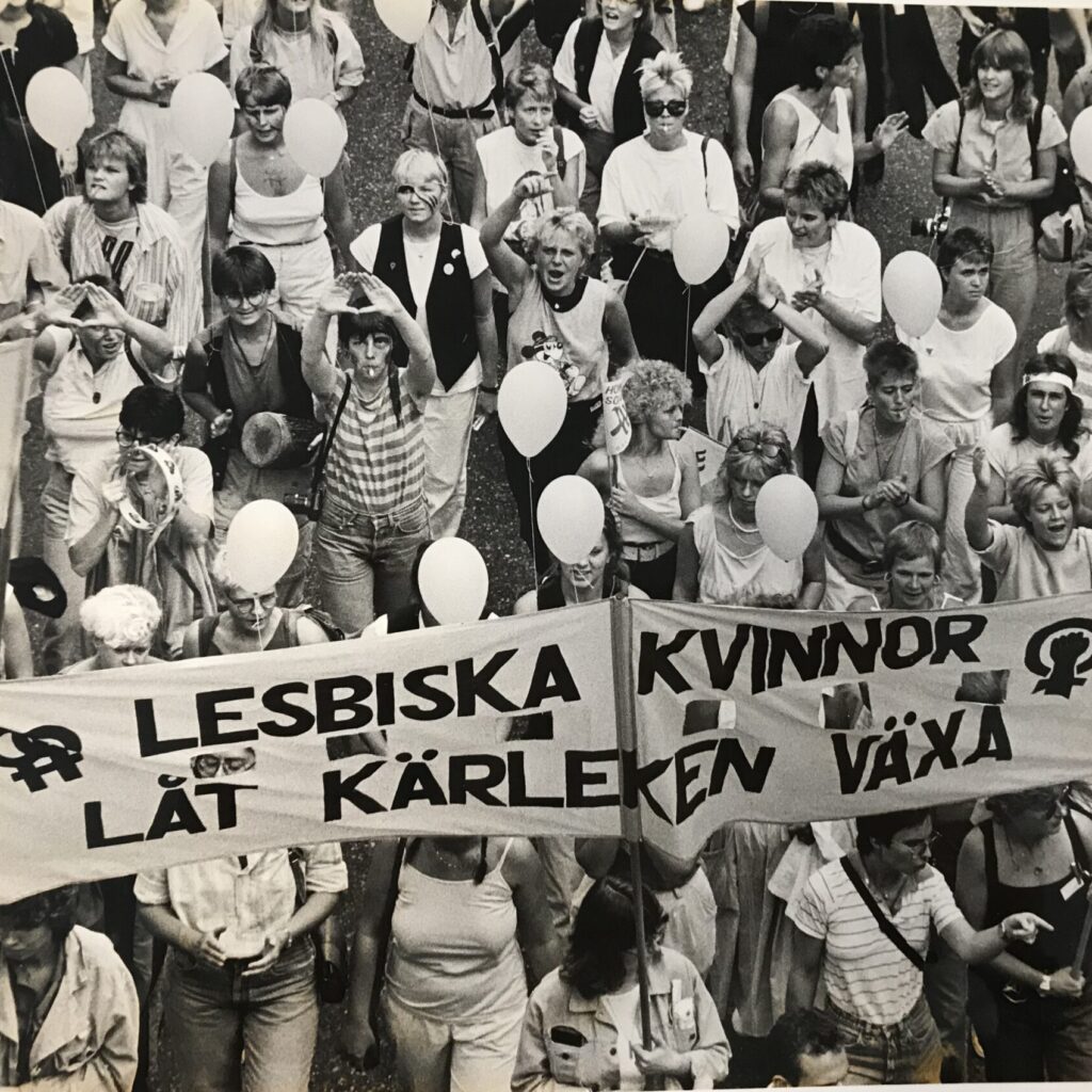 Svart-vitt foto på människor som går i en Prideparad. En banderoll syns i bilden: Lesbiska kvinnor - låt kärleken växa